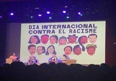 A group of people sitting on a stage in front of a screen that says dia internacional contra el racismo.