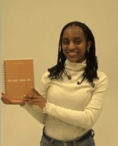 A young woman holding up a notebook in front of a white wall.