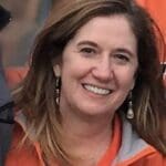 Smiling woman with brown hair wearing orange jacket.