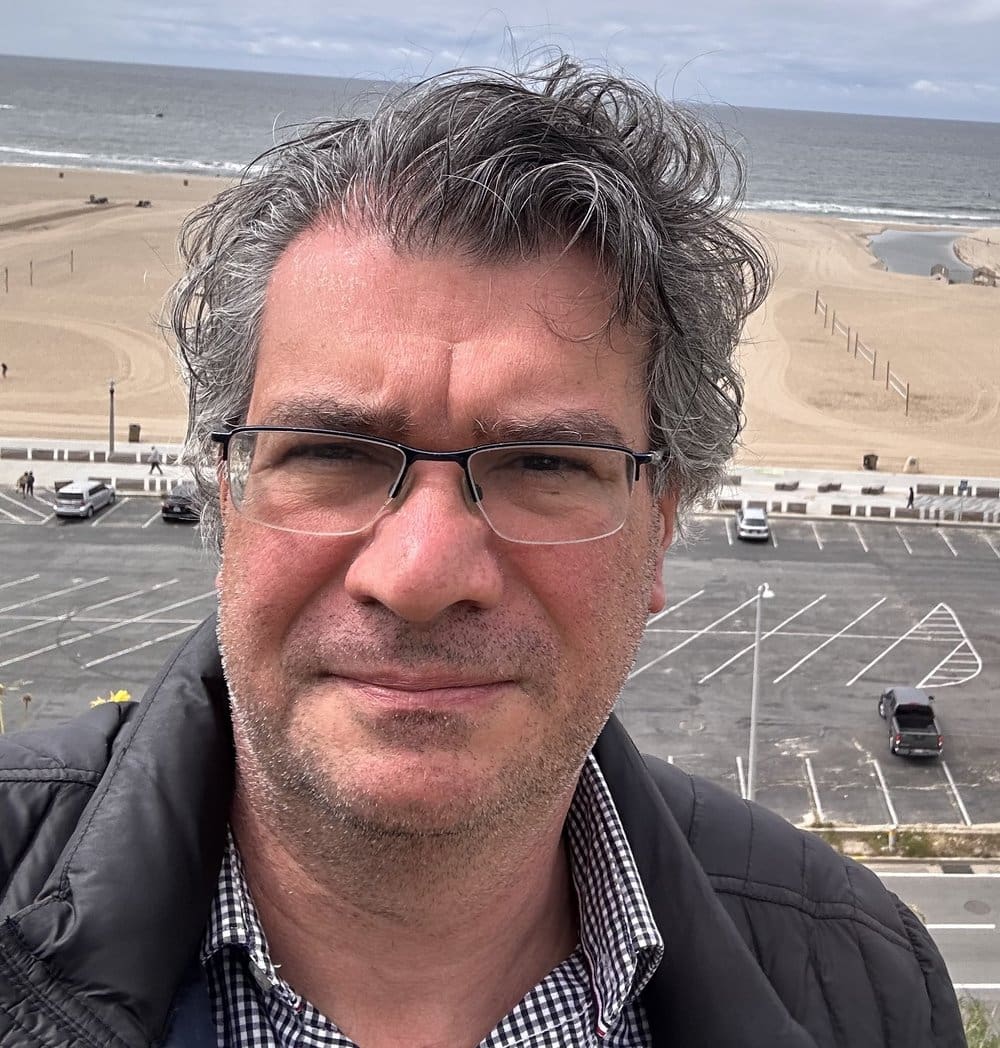 Man wearing glasses with beach in background.