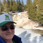 Man in sunglasses by a snowy waterfall.